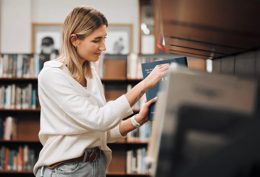 Studentka wybiera książkę z półki w bibliotece.