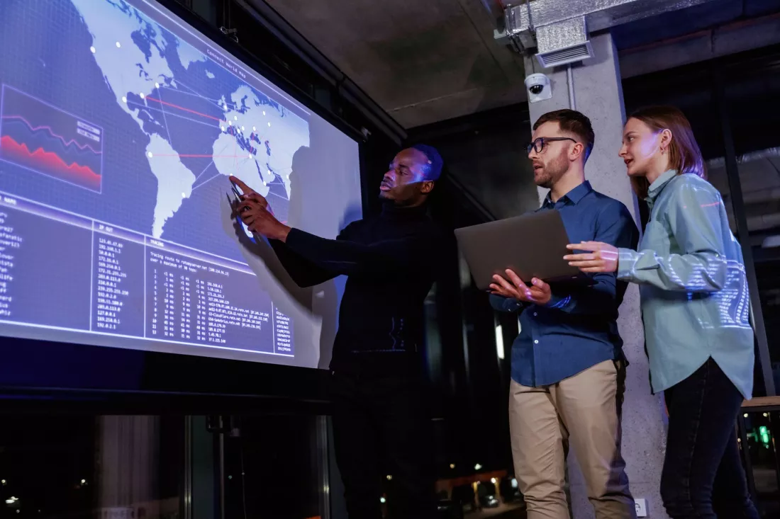 Young analysts discuss data on a large digital world map in a dark operations room