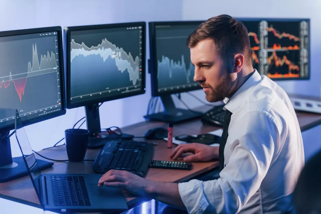 Financial analyst working at a desk with several monitors showing market charts