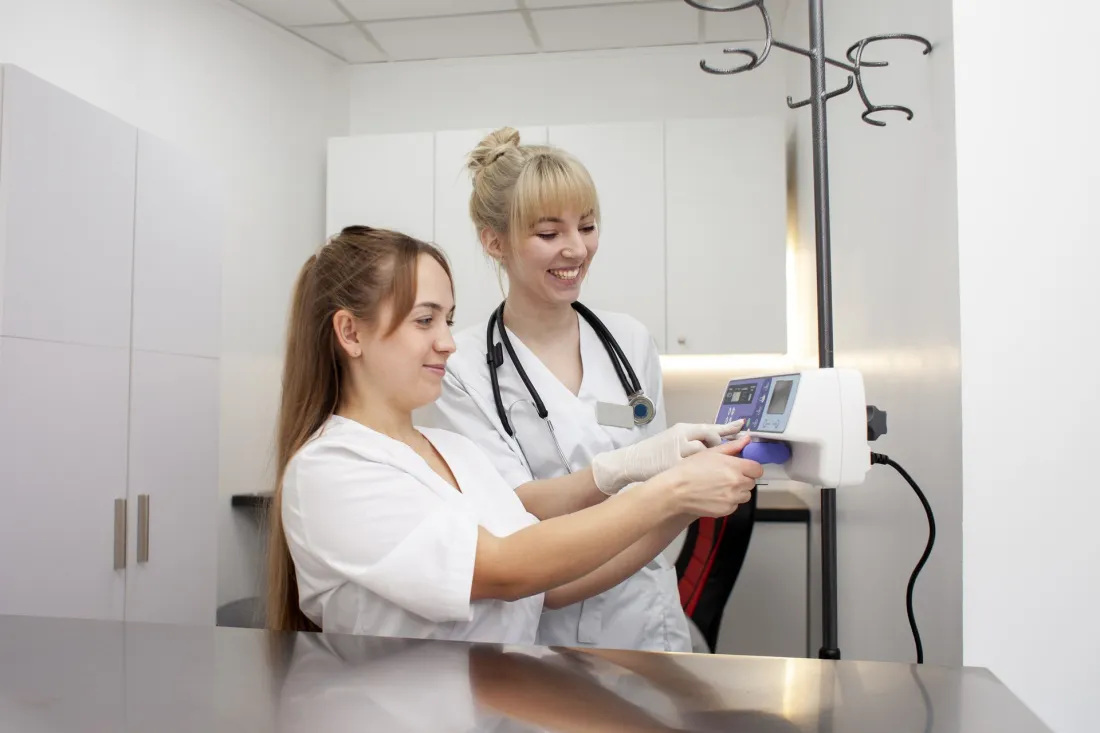 Two young nurses in white uniforms set up an infusion pump on a drip stand in a hospital room.